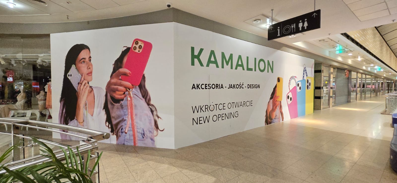 Wnętrze galerii handlowej z reklamą sklepu Kamalion z akcesoriami GSM. Grafika przedstawia osoby z kolorowymi etui na telefony. Napis: Akcesoria - Jakość - Design, Wkrótce otwarcie.