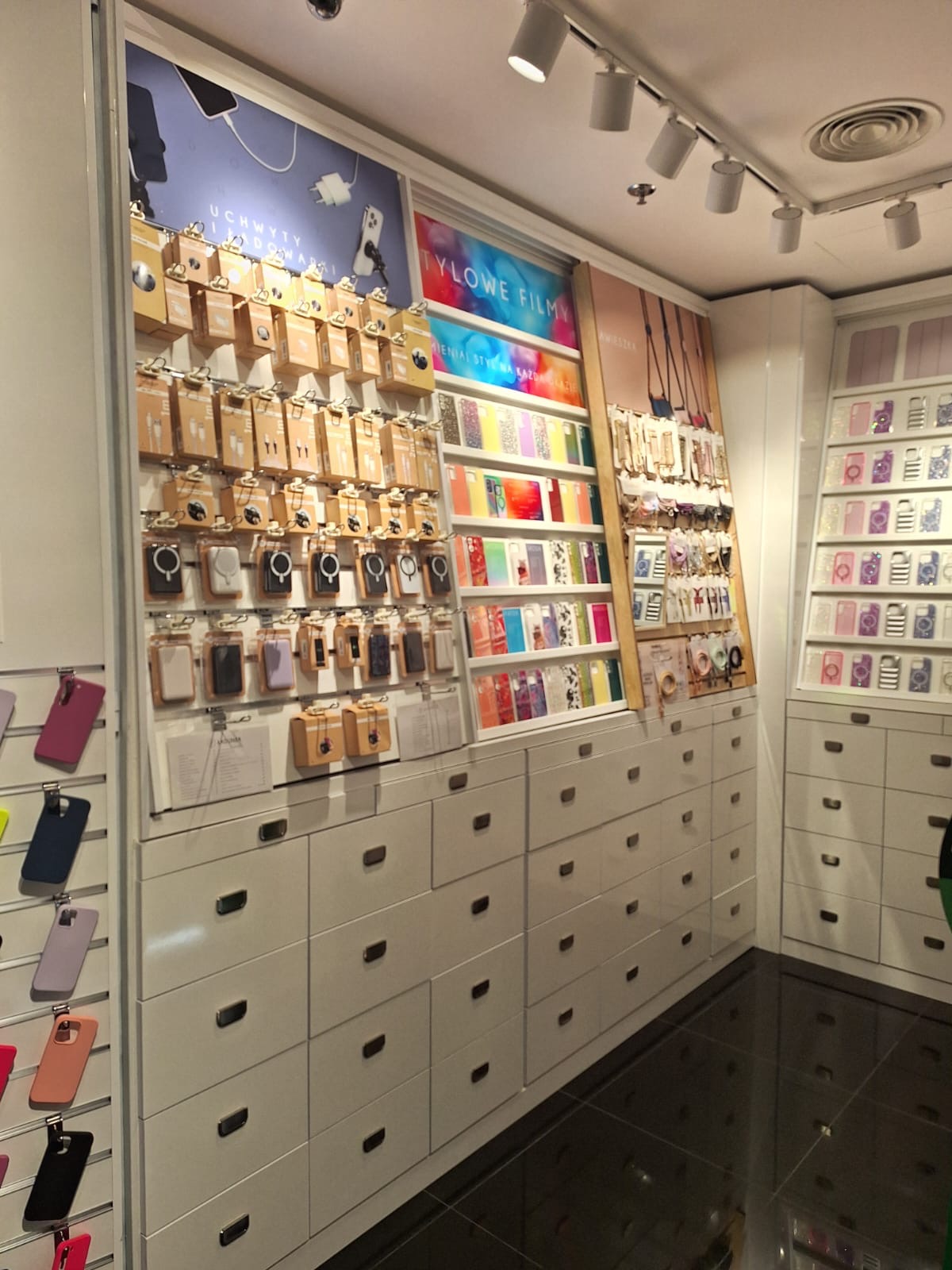Ekspozycja akcesoriów GSM: etui, folie, uchwyty. Różnorodność wzorów i kolorów na białych regałach z szufladami. Oświetlenie punktowe. Widok z perspektywy klienta.