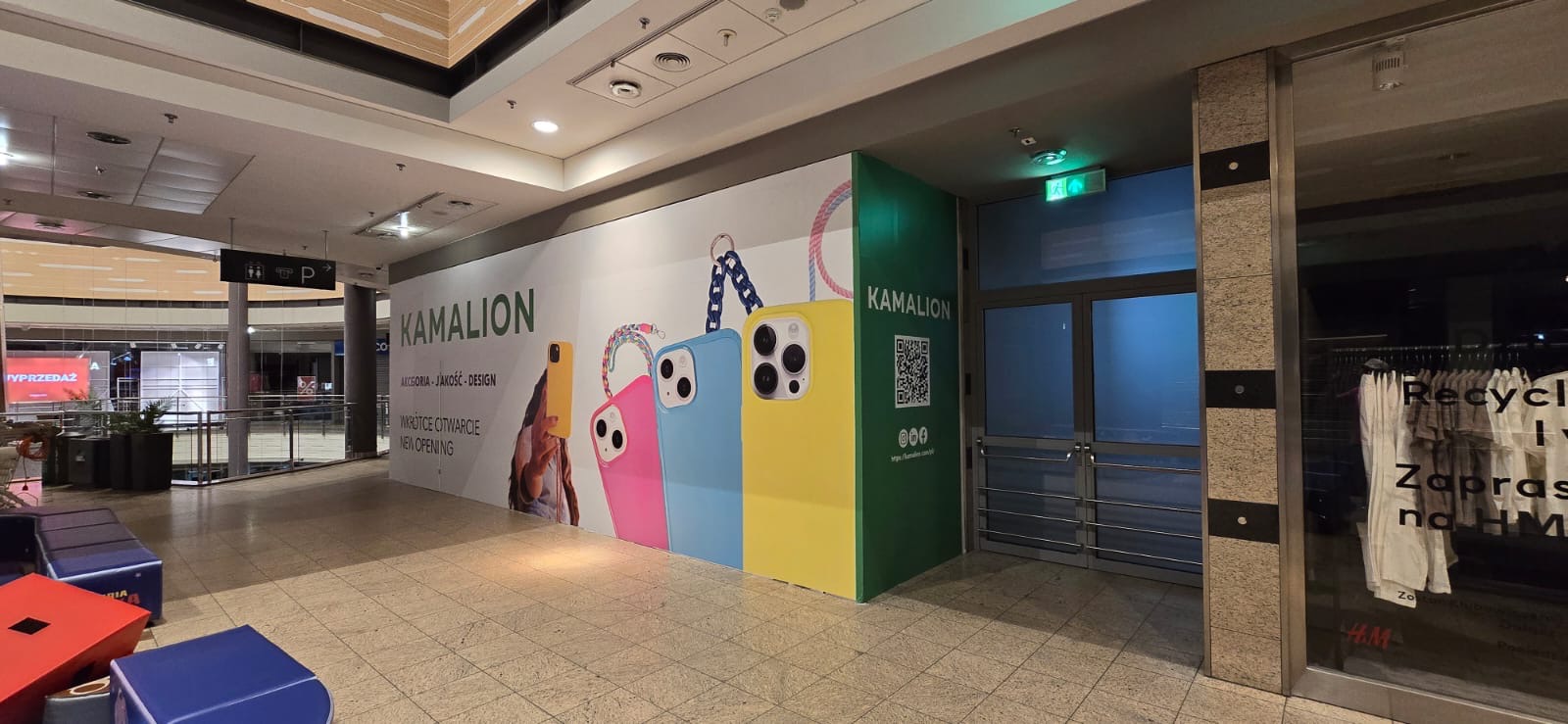 Wnętrze galerii handlowej z reklamą sklepu Kamalion, prezentującą kolorowe etui na telefony i informację o otwarciu. Widoczny kod QR i logo sklepu.