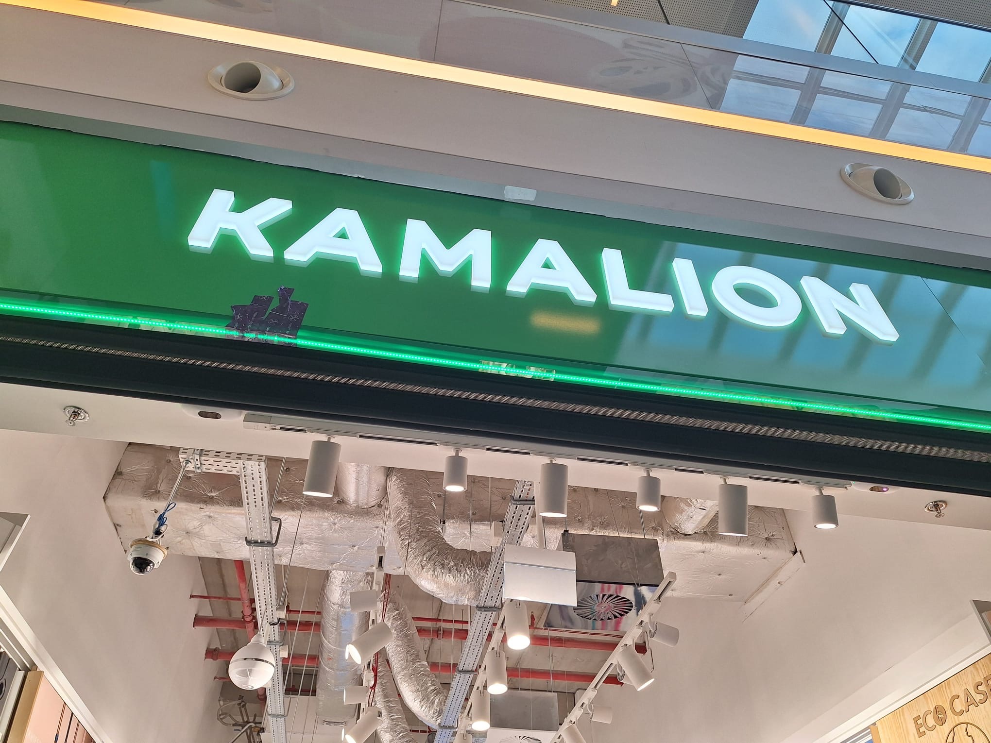 Świecący szyld z nazwą 'KAMALION' na zielonym tle, podświetlony zielonymi diodami LED. Widoczne elementy instalacji sufitowej i oświetlenie punktowe.