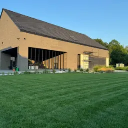 Nowoczesny dom z brązowej cegły z idealnie przystrzyżonym trawnikiem w paski, dekoracyjne kamienie i rośliny ozdobne przy fundamencie, rusztowanie przy oknie.