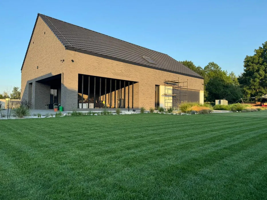 Nowoczesny dom z brązowej cegły z idealnie przystrzyżonym trawnikiem w paski, dekoracyjne kamienie i rośliny ozdobne przy fundamencie, rusztowanie przy oknie.