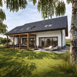 Biuro Projektowe Julia Szatko - Nowoczesny dom z ciemną dachówką i drewnianą pergolą, otoczony zielonym trawnikiem i brzozami. Widoczne leżaki i meble ogrodowe. Ujęcie w słoneczny dzień.