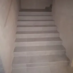 Schody wykończone jasnoszarymi płytkami, prowadzące w górę, ściany pokryte gładzią w odcieniach szarości, widoczne niedoskonałości wykończenia na styku płytek i ściany.