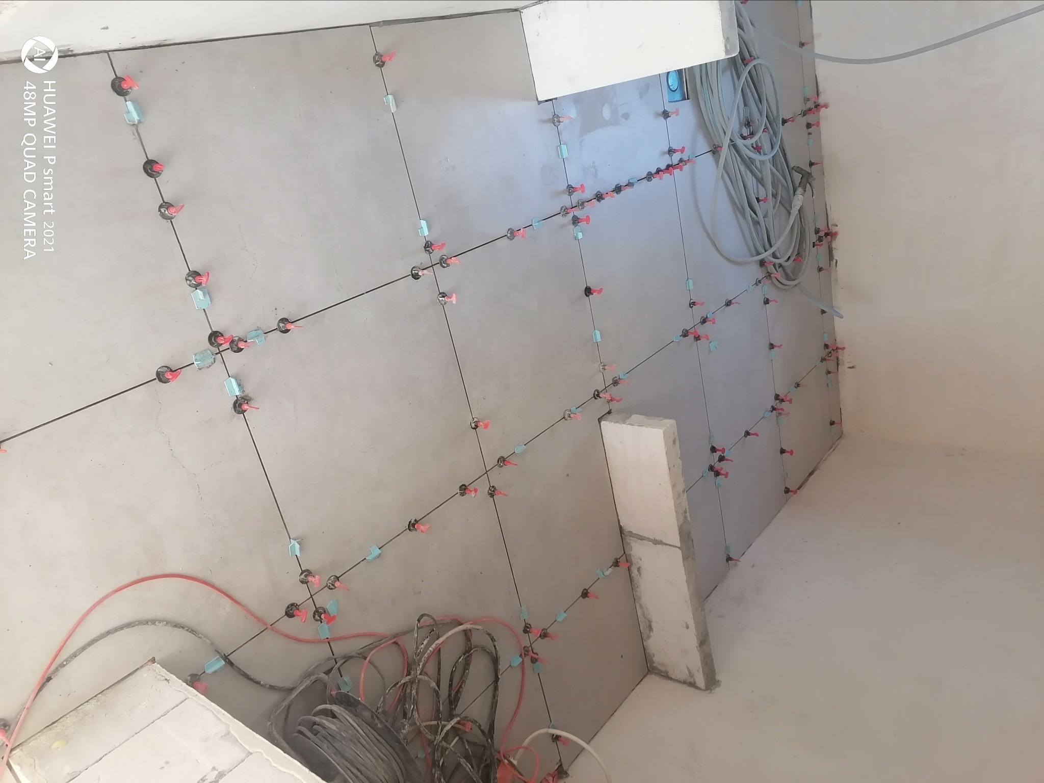 Świeżo położone szare płyty kartonowo-gipsowe na ścianie, z widocznymi systemami poziomowania płytek (klipsy i kliny), oraz zwisającymi kablami elektrycznymi.