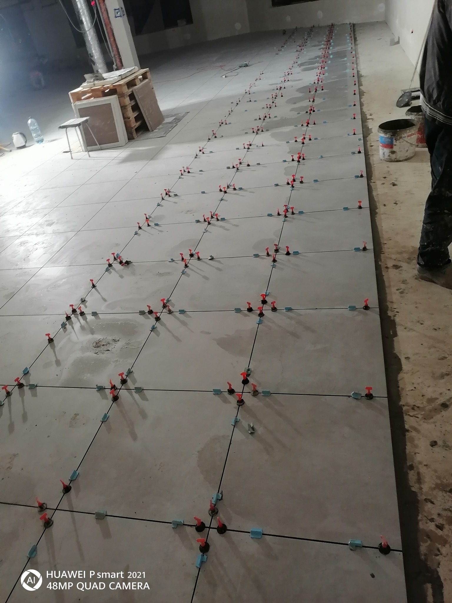 Układanie szarych płytek podłogowych z użyciem systemu poziomowania, widoczne krzyżyki dystansowe i kliny, w tle paleta z materiałami i wiadro.