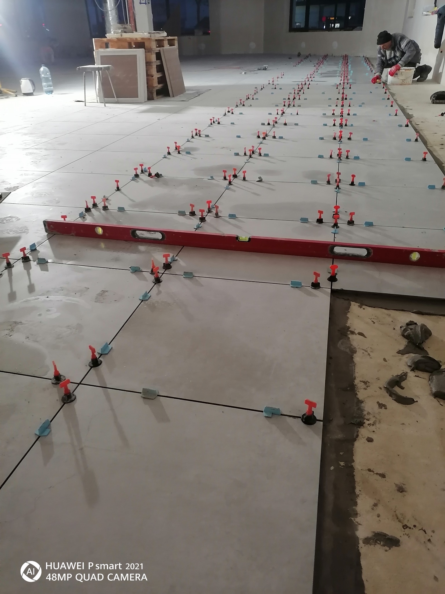 Układanie jasnych płytek podłogowych z wykorzystaniem systemu poziomowania z czerwonymi klinami i niebieskimi dystansami; w tle widoczny pracownik w czapce i roboczych rękawicach.