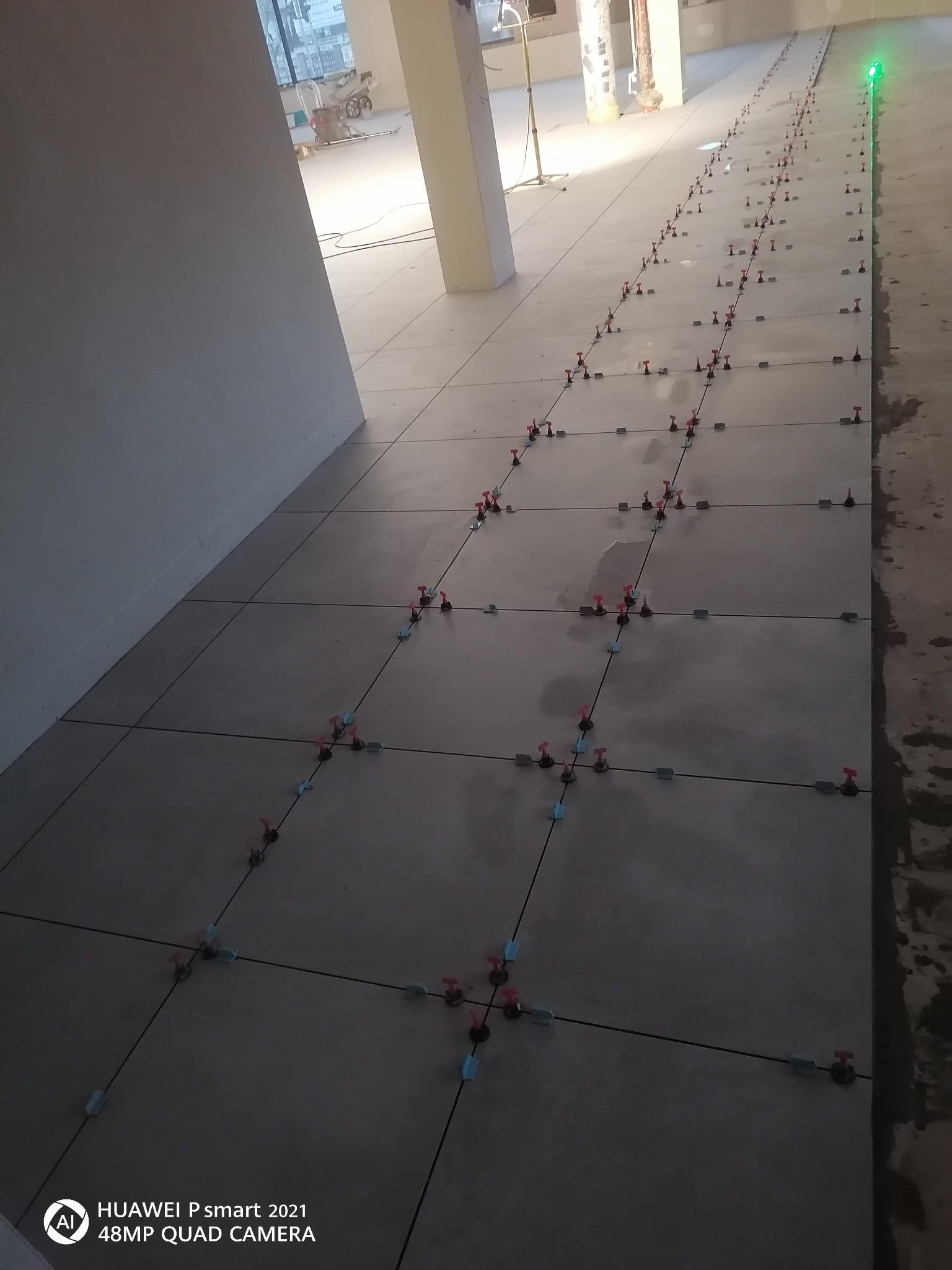 Szare płytki podłogowe w trakcie układania, z widocznymi krzyżykami dystansowymi i systemem poziomowania, oświetlone zielonym laserem poziomującym.