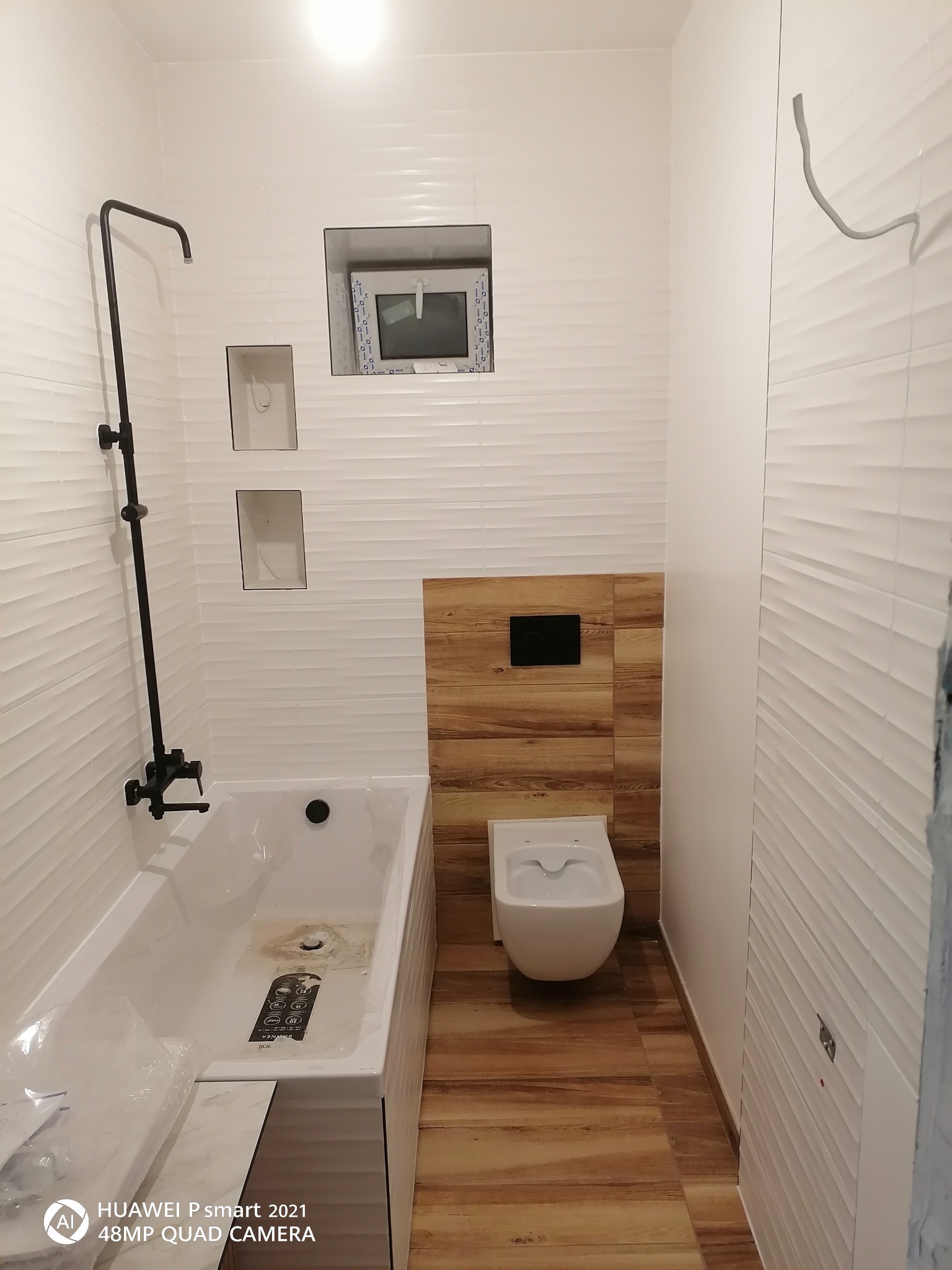 Nowoczesna łazienka z białą armaturą, teksturalnymi białymi płytkami na ścianach, drewnopodobną podłogą i ścianą, czarną baterią prysznicową i wnękami w ścianie