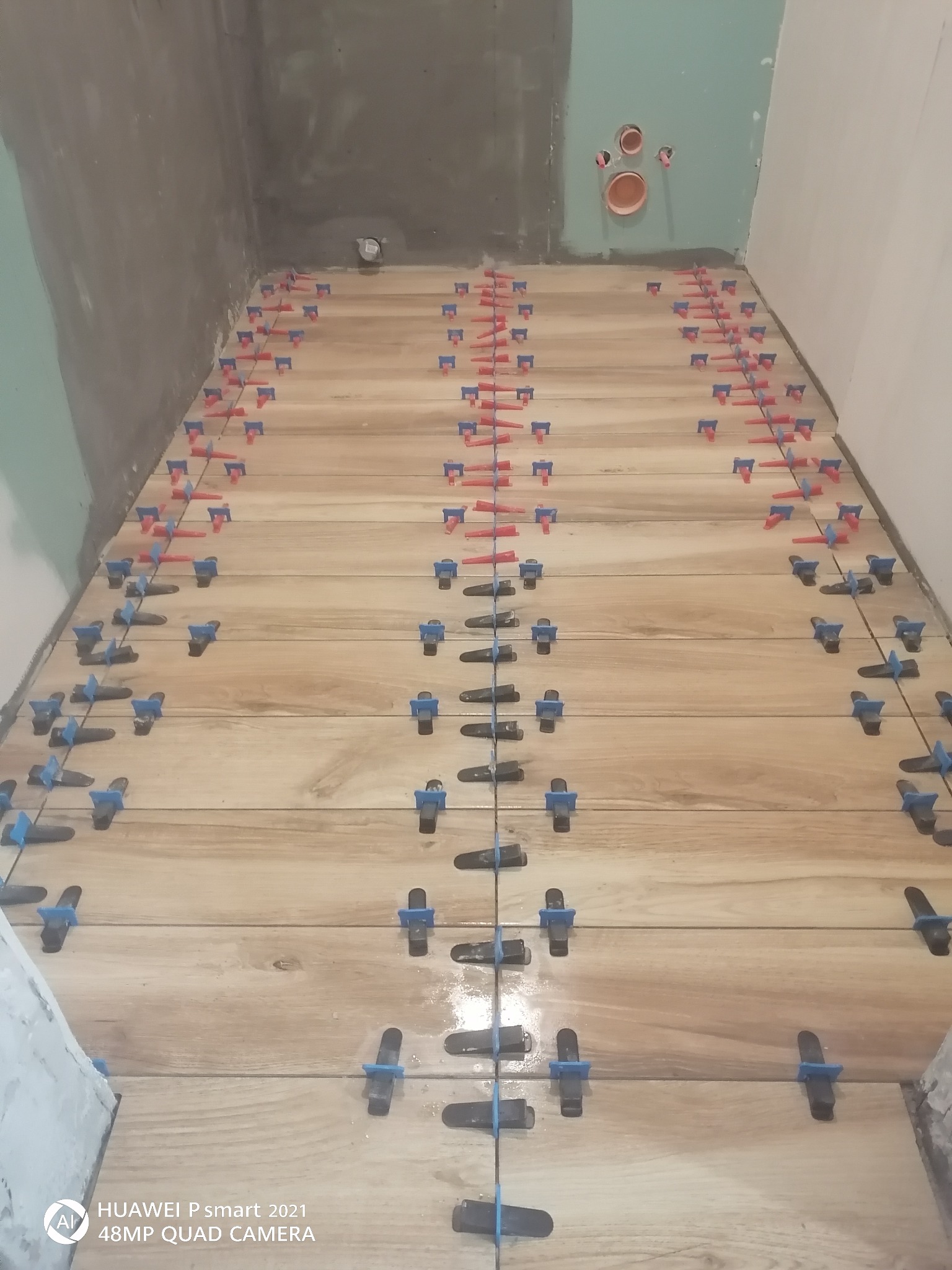 Układanie podłogi z płytek imitujących drewno, z użyciem systemu poziomowania płytek (czerwone i niebieskie klipsy), widoczne niedokończone ściany w tle z wystającymi rurami.