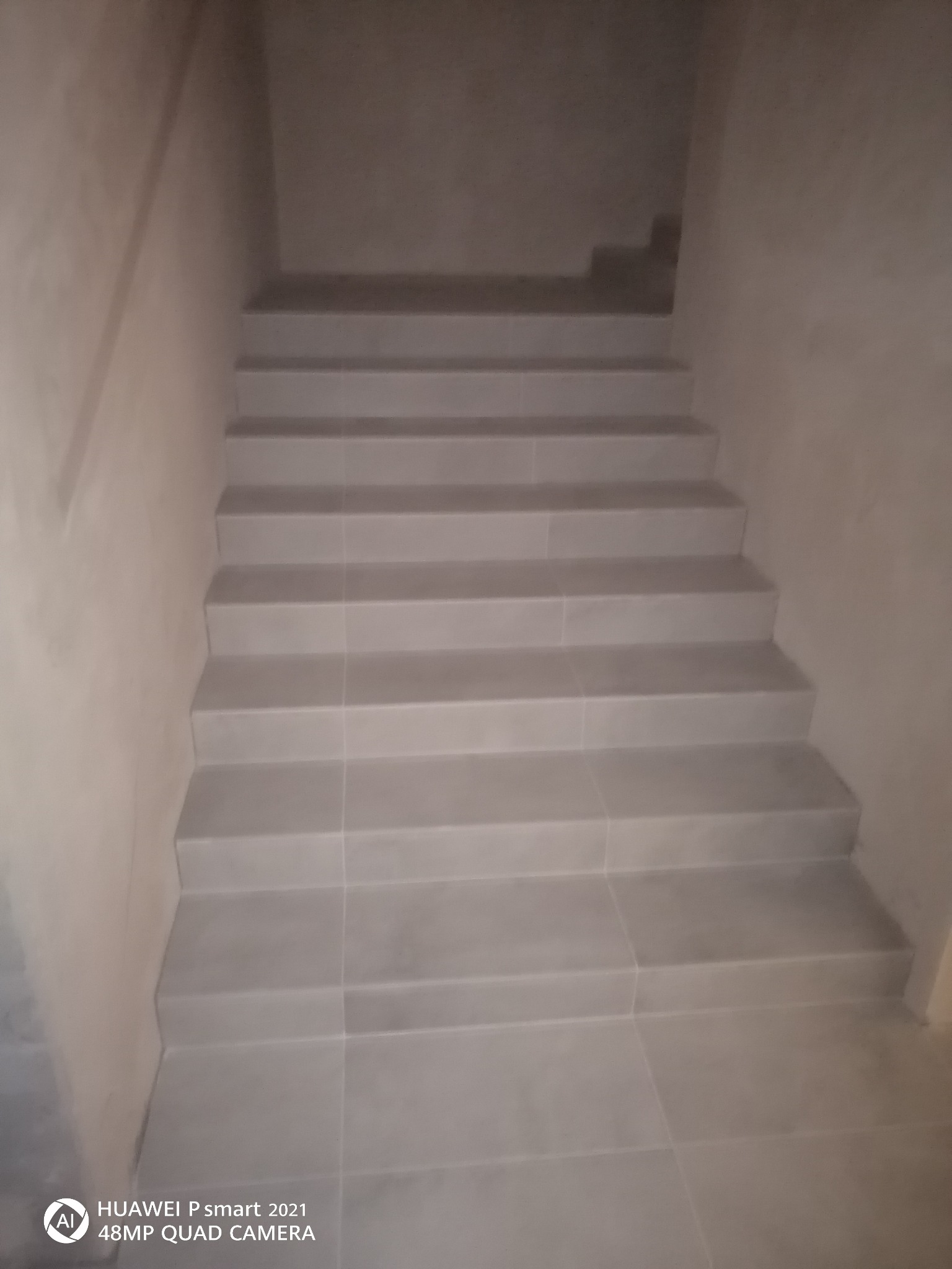 Schody wykończone jasnoszarymi płytkami, prowadzące w górę, ściany pokryte gładzią w odcieniach szarości, widoczne niedoskonałości wykończenia na styku płytek i ściany.