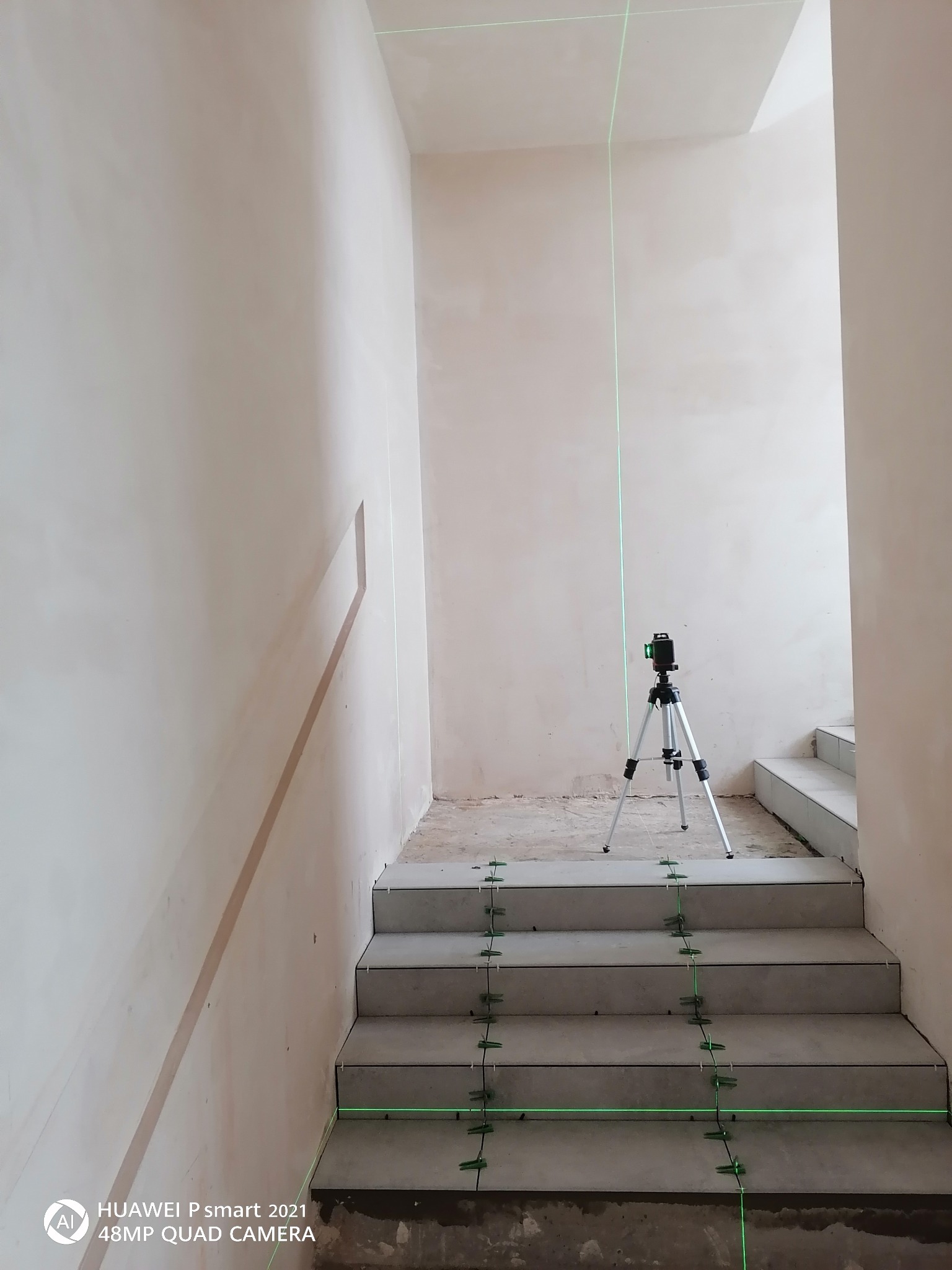 Klatka schodowa w trakcie układania płytek, widoczny laser krzyżowy wyznaczający poziom, plastikowe krzyżyki dystansowe między płytkami.