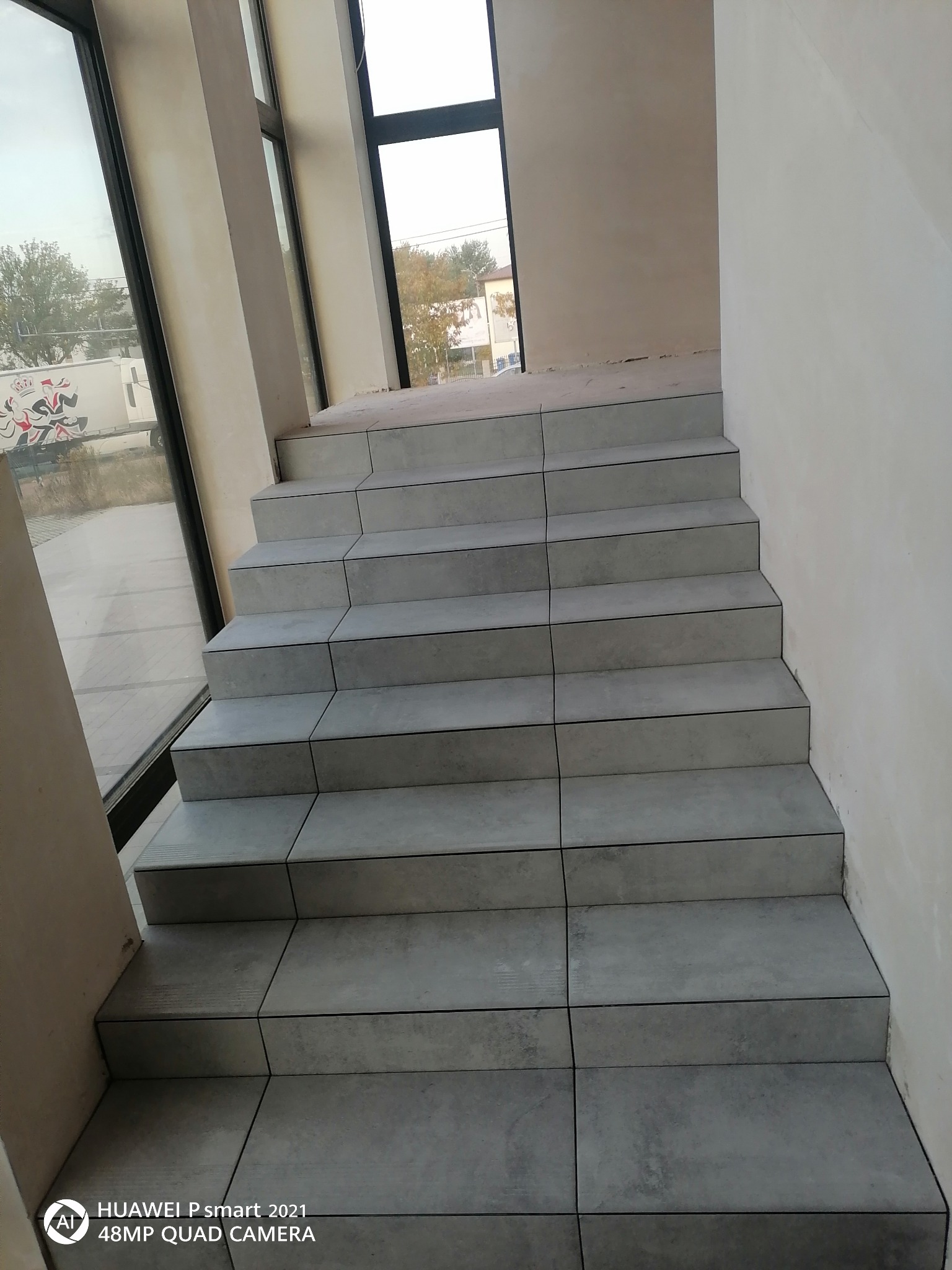 Schody wyłożone szarymi płytkami, widok z dołu, ściany w trakcie wykańczania, duże okno z widokiem na zewnątrz.