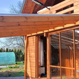 Drewniana konstrukcja zadaszenia tarasu, połączona z budynkiem. Widoczne przesuwne drzwi szklane i drewniane elementy wykończeniowe. W tle tunel foliowy i kwitnące drzewo.