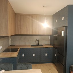 SimWood Szymon Niezgoda - Nowoczesna kuchnia w trakcie montażu: drewniane blaty, niebieskie szafki z folią ochronną, czarna armatura i sprzęt AGD. Widoczne elementy wykończeniowe.