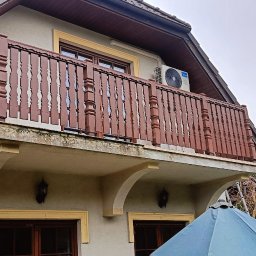 SimWood Szymon Niezgoda - Balkon z brązową, drewnianą balustradą o dekoracyjnych wycięciach, przymocowaną do beżowej elewacji domu. Widoczny klimatyzator na ścianie i fragment niebieskiego parasola.