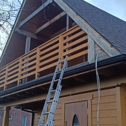 SimWood Szymon Niezgoda - Drewniany dom z balkonem w trakcie budowy, widoczna drabina oparta o ścianę. Balustrada balkonowa z poziomych desek. Dach pokryty gontem bitumicznym.