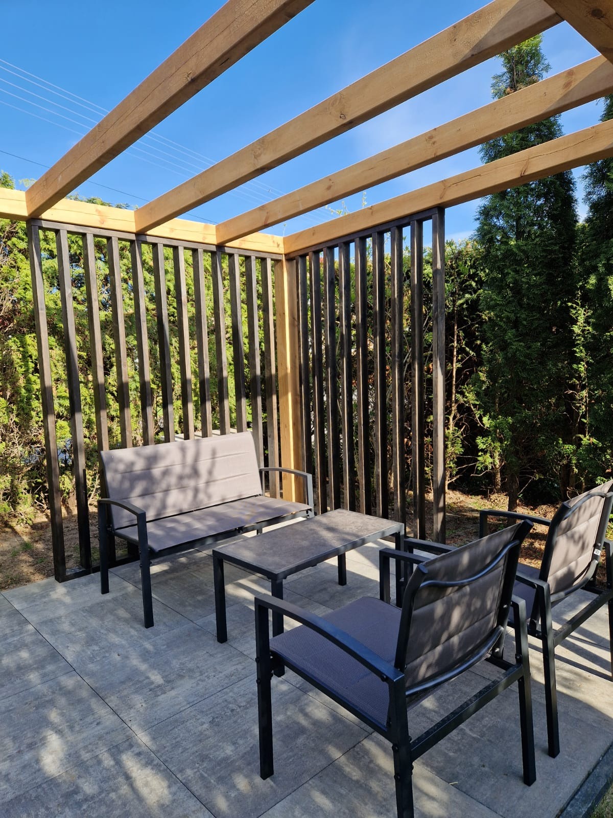 Drewniana pergola z pionowymi listwami w kolorze grafitowym, wyposażona w grafitowy zestaw mebli ogrodowych na szarym tarasie. Widok na zielone tuje i błękitne niebo.