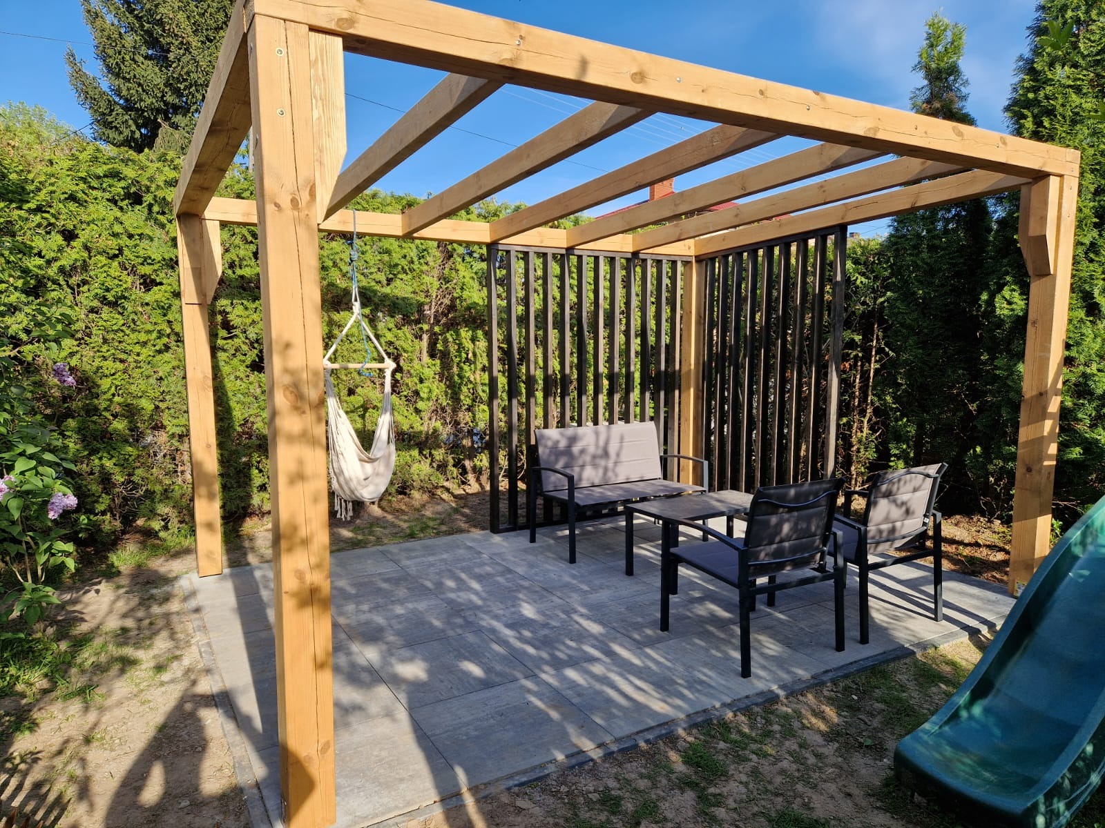 Drewniana pergola z hamakiem i meblami ogrodowymi na brukowanym tarasie, otoczona zielenią. Elementy metalowe w tle. Słoneczny dzień.