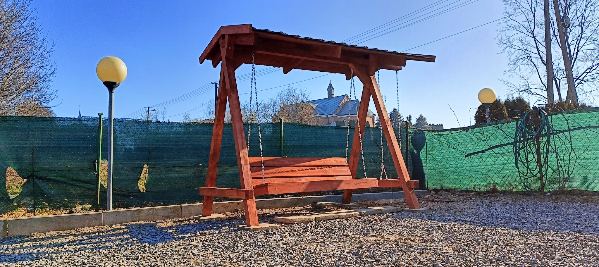 Drewniana huśtawka ogrodowa z zadaszeniem, zawieszona na łańcuchach, z widokiem na kościół w tle, oświetlona słońcem. Otoczenie to żwir i zielona siatka.