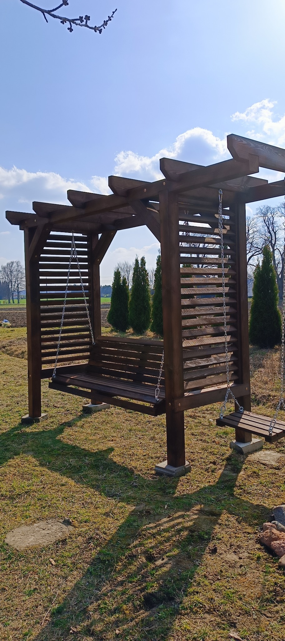 Ciemnobrązowa, drewniana pergola z huśtawką na łańcuchach, ustawiona na trawniku. W tle widoczne drzewa i błękitne niebo z chmurami.