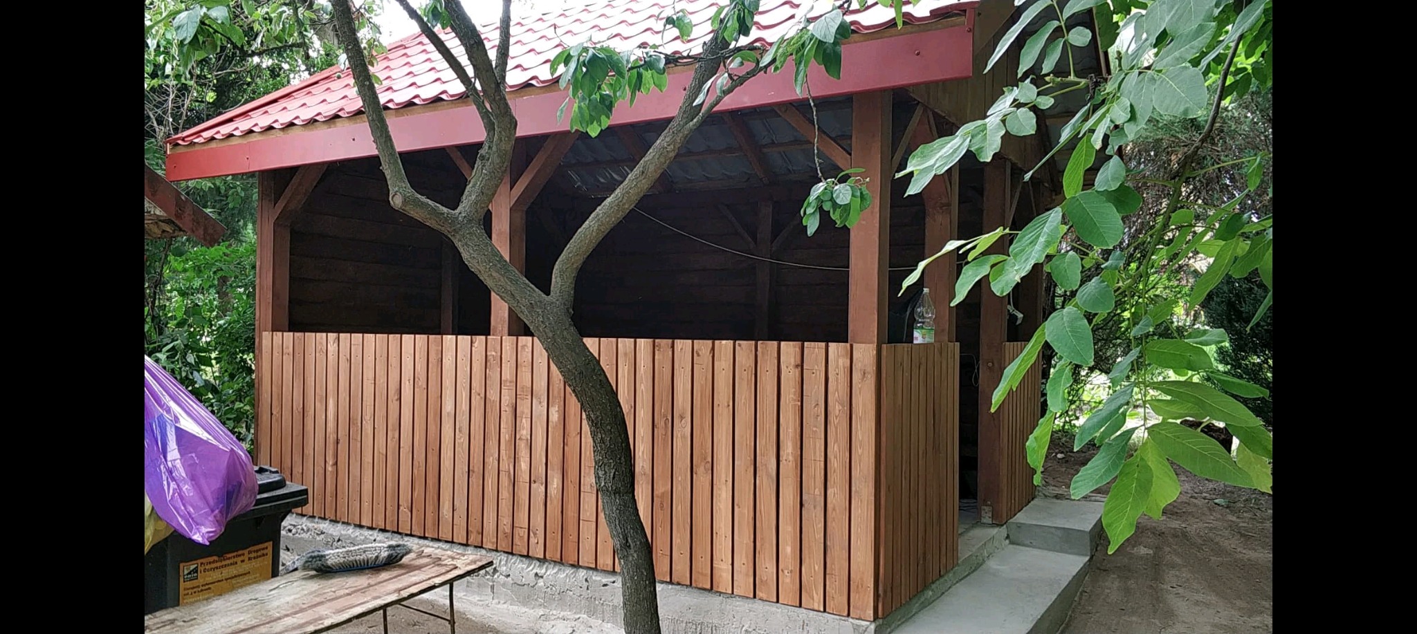 Częściowo zabudowana, drewniana altana z czerwonym dachem, widoczna zza drzewa. Ściany altany z pionowych desek, drewno w ciepłym odcieniu brązu. W tle zieleń.