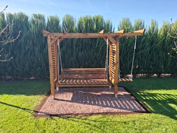 Drewniana huśtawka ogrodowa z pergolą, zawieszona na łańcuchach, na tle zielonego żywopłotu i trawnika. Podłoże wysypane drobnym żwirem. Słoneczny dzień.