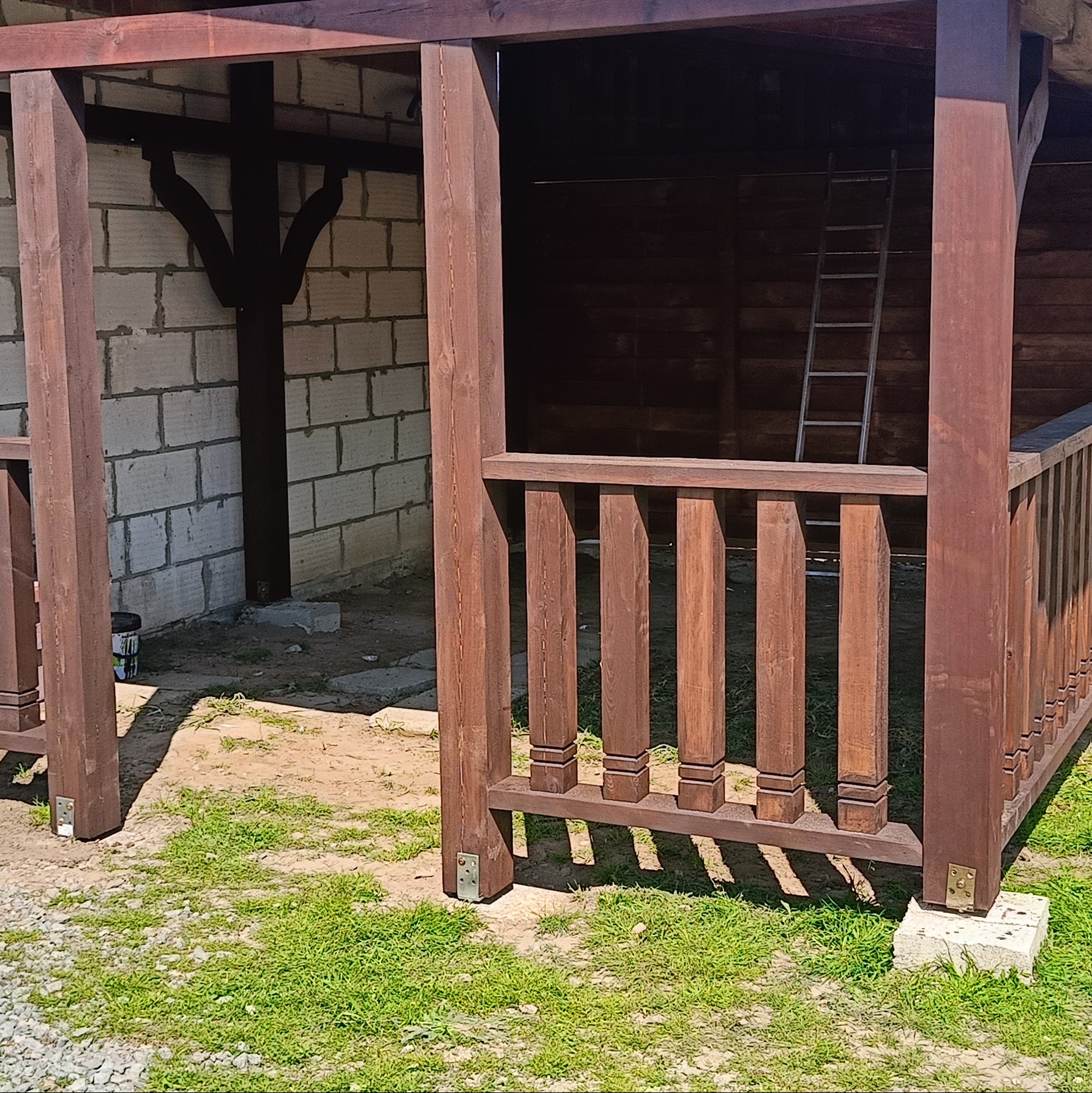 Fragment konstrukcji altany z brązowego drewna, z widoczną balustradą i elementami wspornymi. W tle ściana z pustaków i drabina.