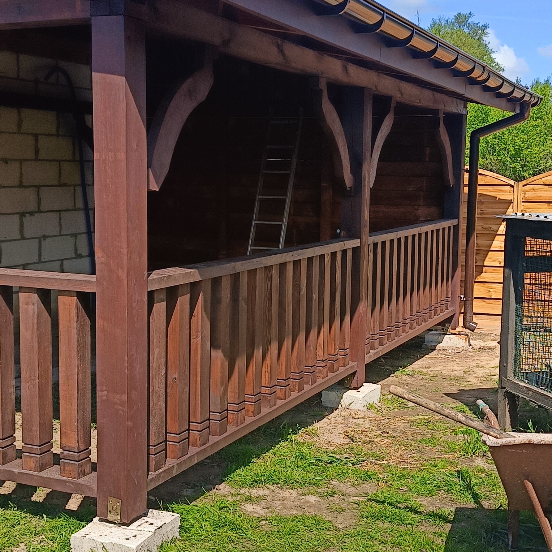 Zadaszona, brązowa konstrukcja z drewnianą balustradą, wsparta na betonowych bloczkach. W tle widoczna drabina oparta o ścianę i zielona roślinność.