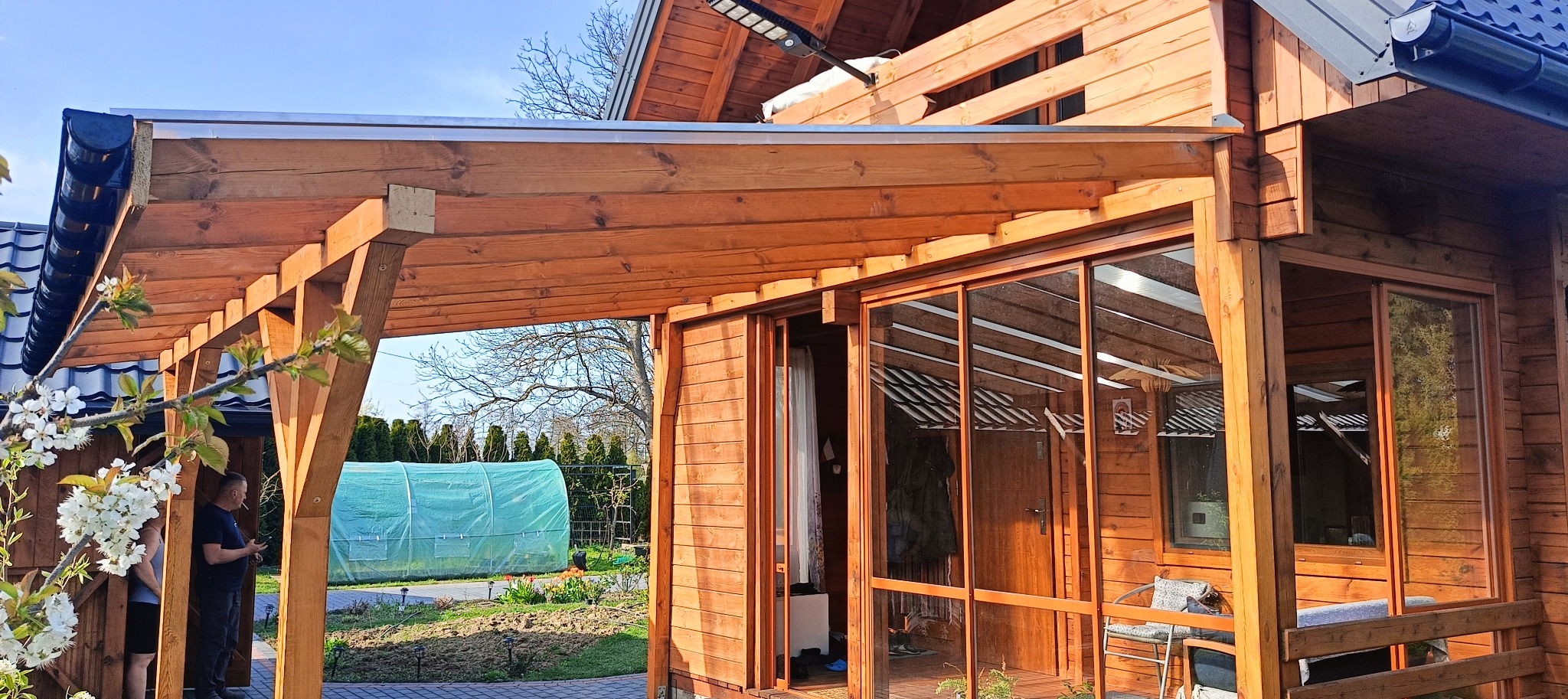 Drewniana konstrukcja zadaszenia tarasu, połączona z budynkiem. Widoczne przesuwne drzwi szklane i drewniane elementy wykończeniowe. W tle tunel foliowy i kwitnące drzewo.