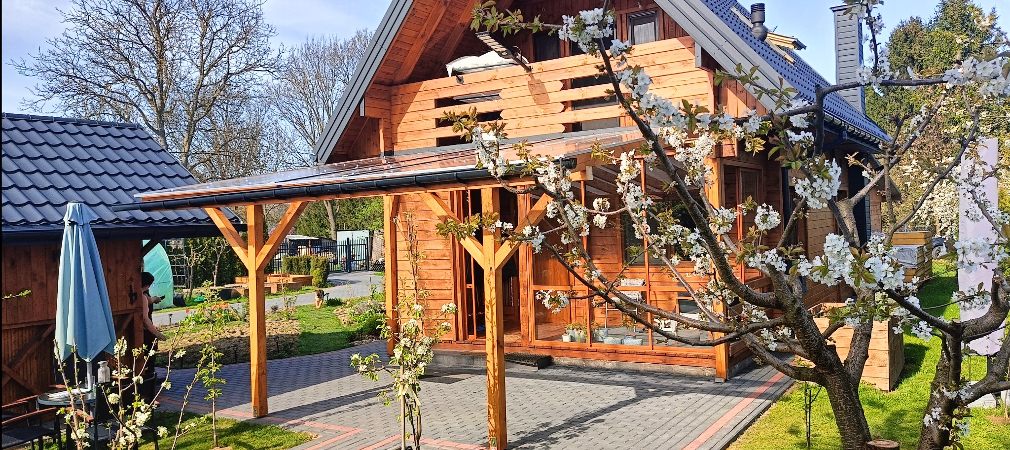 Drewniany dom z przeszkloną werandą i pergolą, otoczony kwitnącymi drzewami owocowymi. Widoczny fragment kostki brukowej i trawnika. Słoneczny dzień.