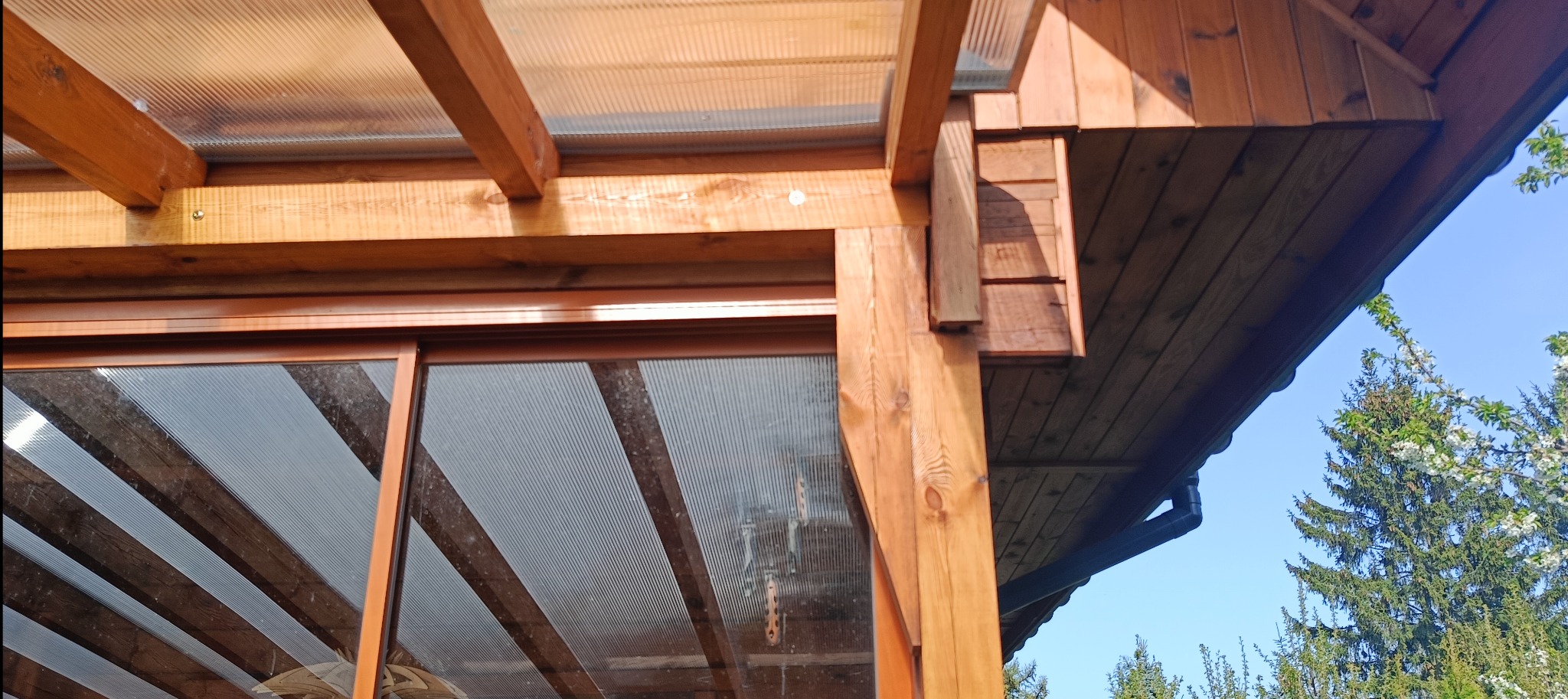 Fragment konstrukcji z drewna i poliwęglanu. Widoczne elementy zadaszenia i ściany, detale połączeń, na zewnątrz zieleń drzew i błękit nieba.