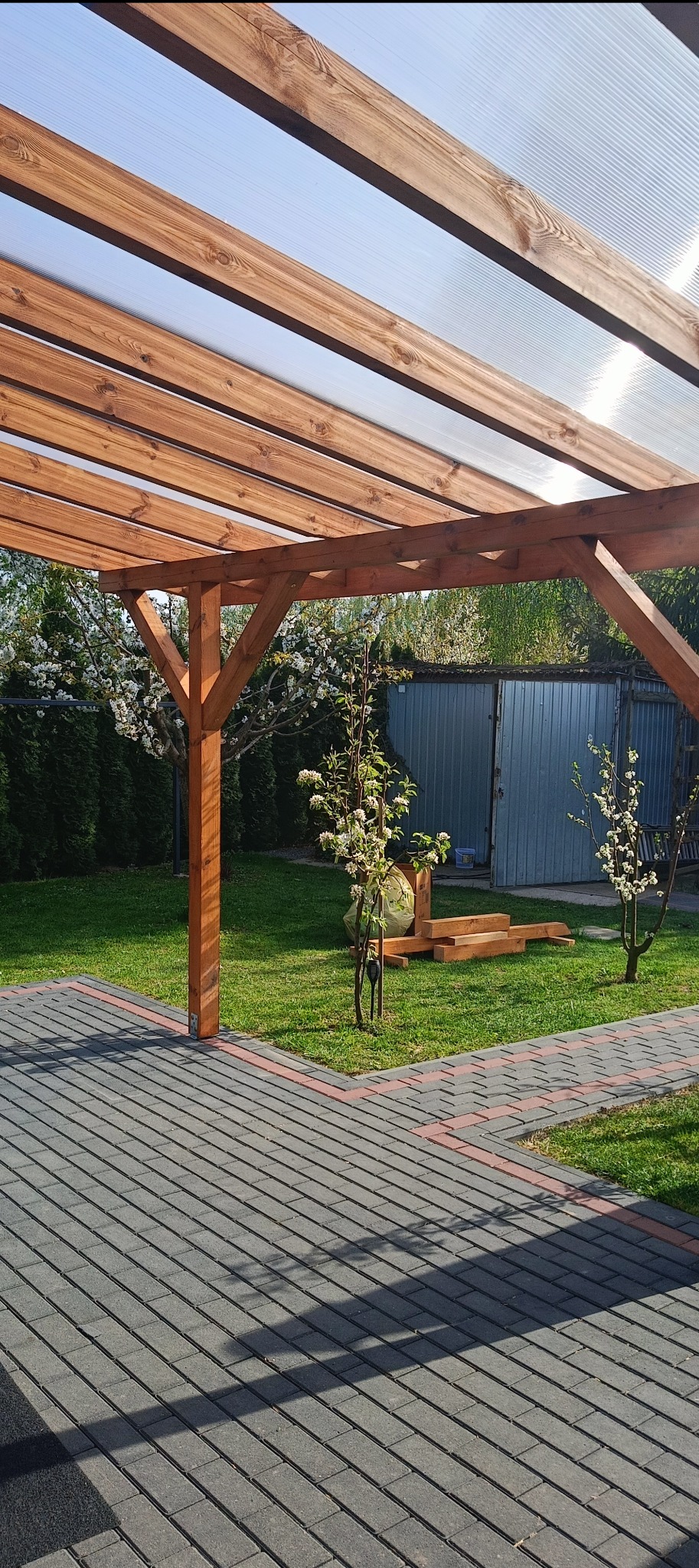 Drewniana pergola z przezroczystym zadaszeniem na tle zielonego ogrodu i kostki brukowej. Widoczne kwitnące drzewa owocowe i elementy konstrukcyjne z drewna.