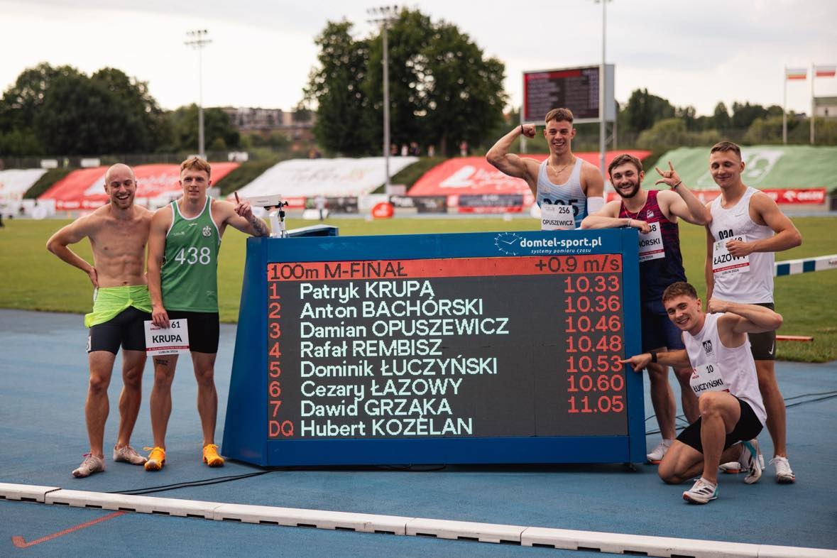 Grupa młodych sportowców pozuje do zdjęcia po finale biegu na 100m, prezentując wyniki na tablicy świetlnej. Widoczne numery startowe, stroje sportowe i uśmiechnięte twarze na tle stadionu.