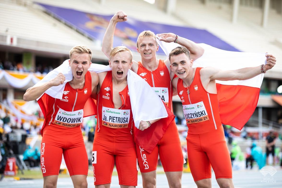 Czterech sportowców w czerwonych strojach reprezentacji Polski świętuje zwycięstwo, owinięci biało-czerwoną flagą na tle stadionu w Nairobi.