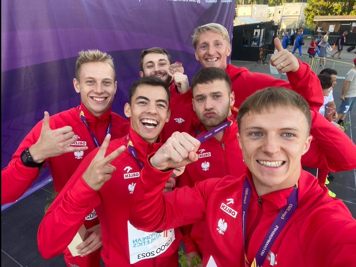 Grupa uśmiechniętych sportowców w czerwonych dresach z godłem Polski i medalami na szyjach, pozujących do selfie po zawodach.