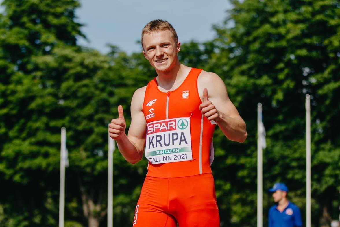 Uśmiechnięty sportowiec w pomarańczowym stroju reprezentacji Polski, z logo sponsorów, stojący na tle zieleni i flag, z uniesionymi kciukami w górę.