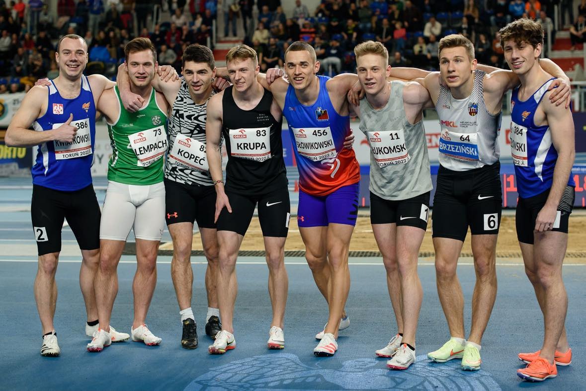 Grupa ośmiu młodych mężczyzn, ubranych w stroje sportowe z numerami startowymi, stoi ramię w ramię na bieżni krytej hali, w tle widownia.