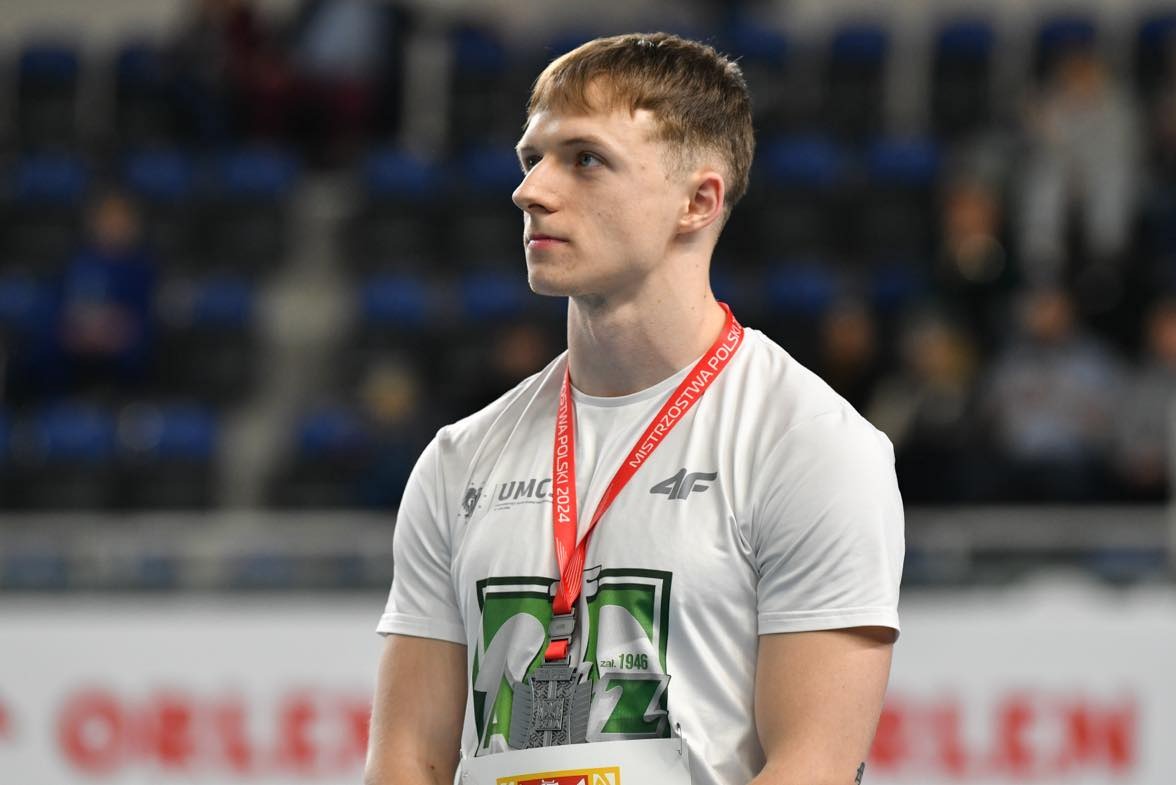 Młody mężczyzna z medalem na szyi, ubrany w białą koszulkę z logo, po zawodach sportowych.