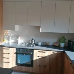 Jasne, nowoczesne meble kuchenne z drewnianymi frontami szafek dolnych i szarymi górnymi. Na blacie ekspres do kawy i przyprawy. W tle okno z promieniami słońca.