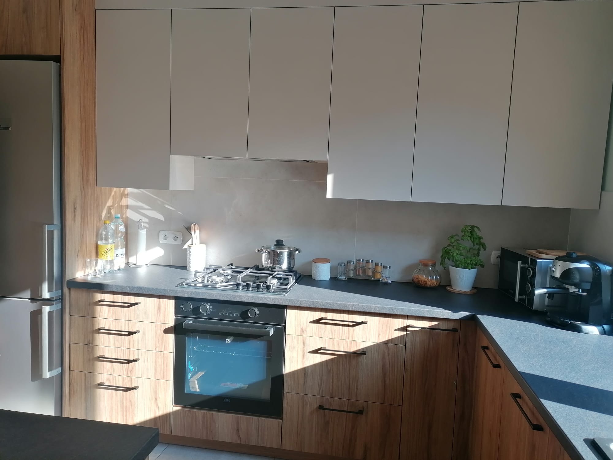 Jasne, nowoczesne meble kuchenne z drewnianymi frontami szafek dolnych i szarymi górnymi. Na blacie ekspres do kawy i przyprawy. W tle okno z promieniami słońca.