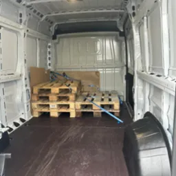 Wnętrze czystego, białego busa transportowego z paletami zabezpieczonymi niebieskimi pasami transportowymi, widoczne ślady użytkowania na podłodze.