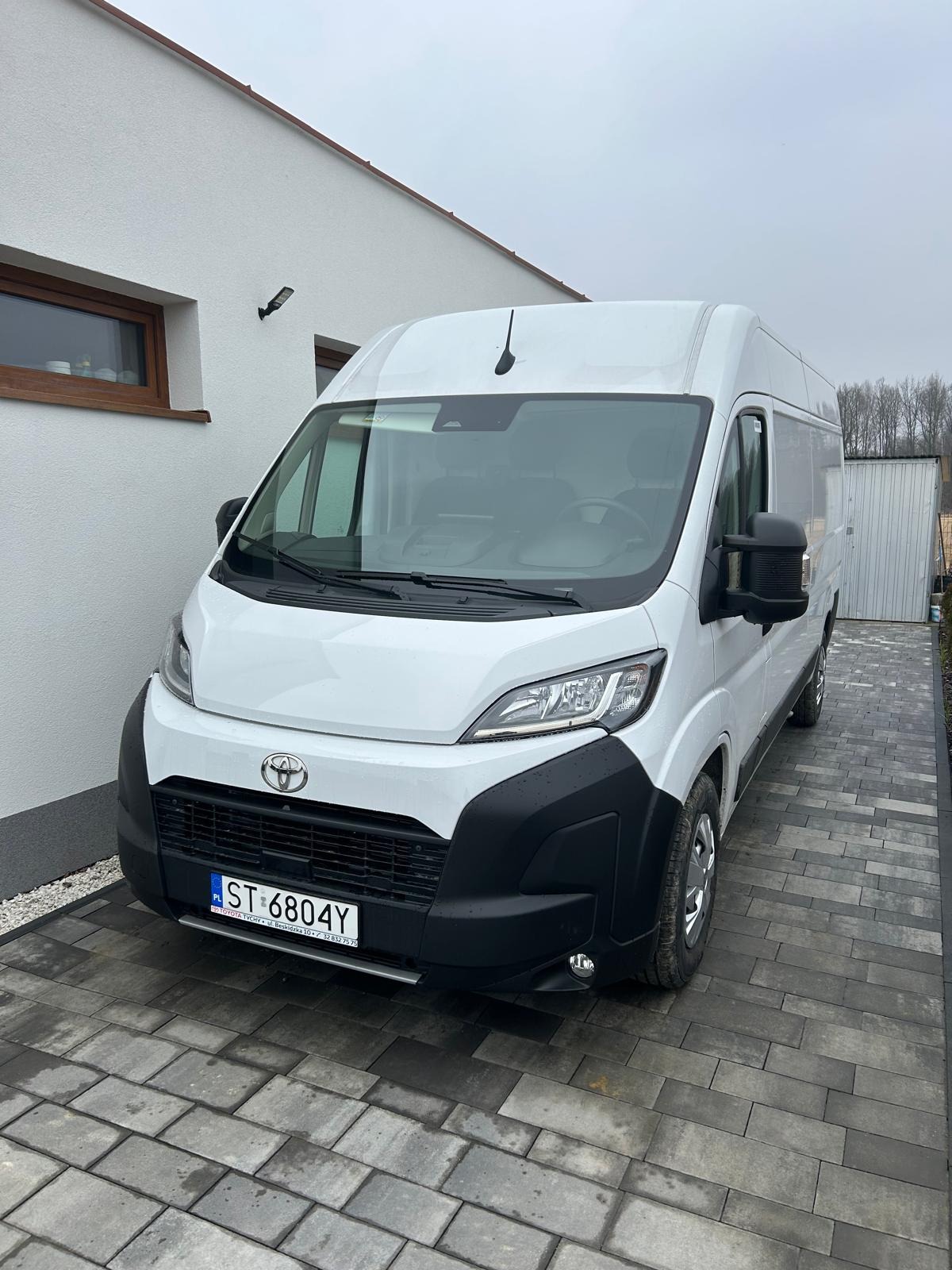 Biały bus marki Toyota zaparkowany na brukowanej posesji przed białym domem z drewnianymi oknami, widoczna tablica rejestracyjna ST 6804Y.