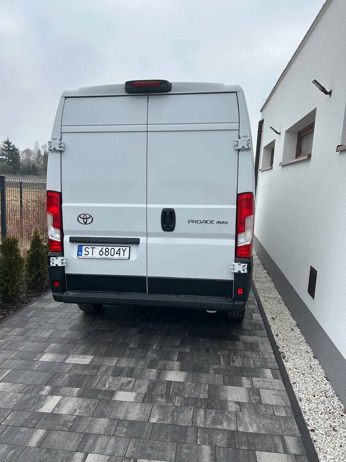 Tył białego busa Toyota Proace Max z otwartymi drzwiami, zaparkowanego na szarej kostce brukowej przed białym budynkiem z małymi oknami.