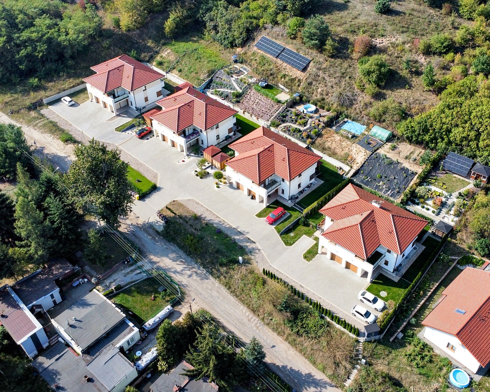 Widok z lotu ptaka na osiedle domów jednorodzinnych z czerwonymi dachami, jasnymi elewacjami, zadbanymi ogrodami i panelami słonecznymi na wzgórzu w tle.