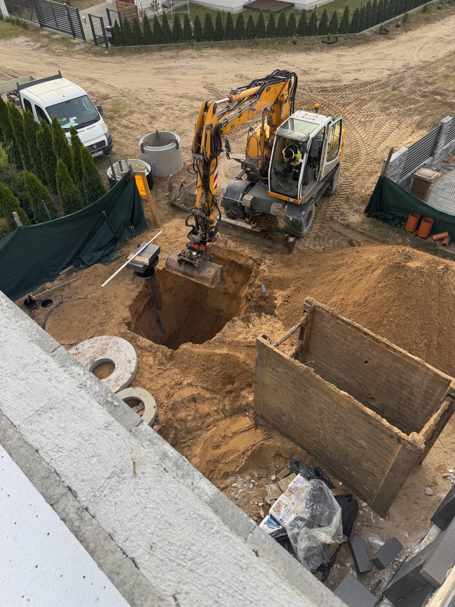 Żółta koparka zagłębia się w wykop w ziemi, obok leżą betonowe kręgi, widoczna szalunkowa skrzynia, w tle zaparkowany biały samochód dostawczy i rząd tuj.