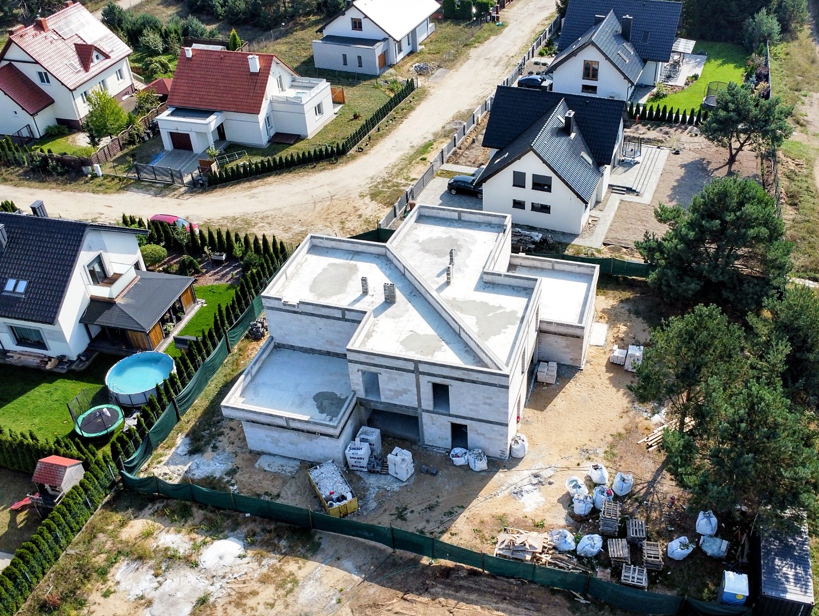 Zdjęcie z lotu ptaka przedstawia budowę domu z betonu komórkowego, widoczne ściany w stanie surowym otwartym, materiały budowlane na działce i sąsiednie zabudowania.