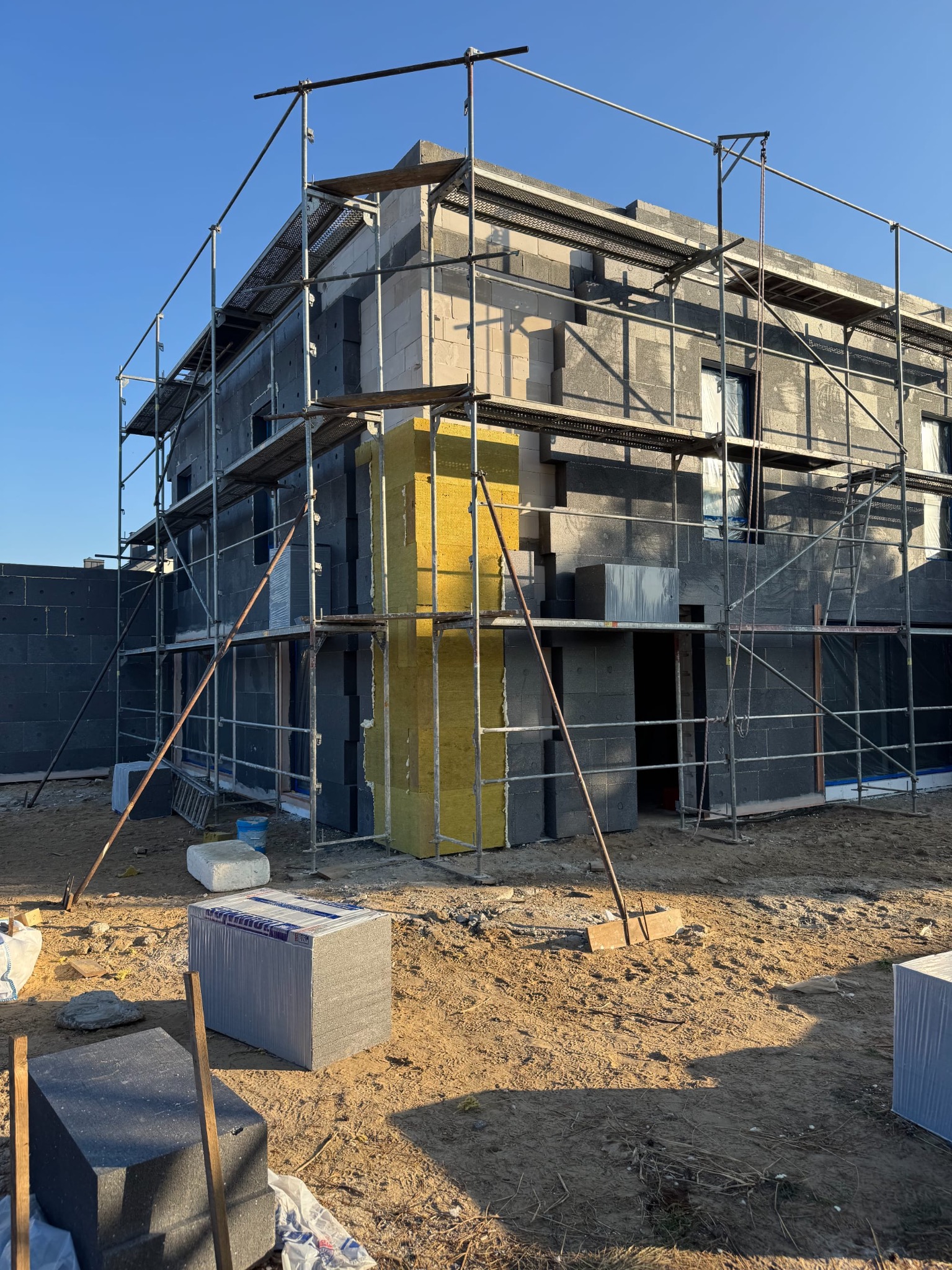 Wznoszony dom z szarego betonu komórkowego, częściowo ocieplony żółtą wełną mineralną, otoczony rusztowaniem w słoneczny dzień.