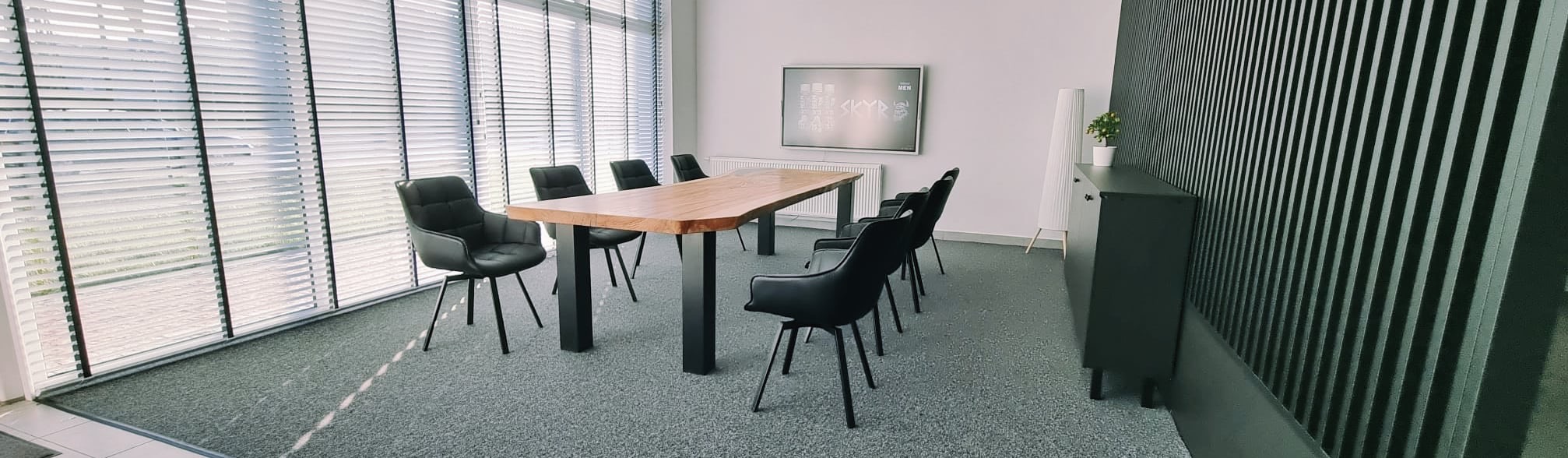 Nowoczesna sala konferencyjna z drewnianym stołem na czarnych nogach, otoczonym przez czarne skórzane krzesła, szare wykładziny dywanowe, żaluzje w oknach i dekoracyjną ścianą z pionowych paneli.