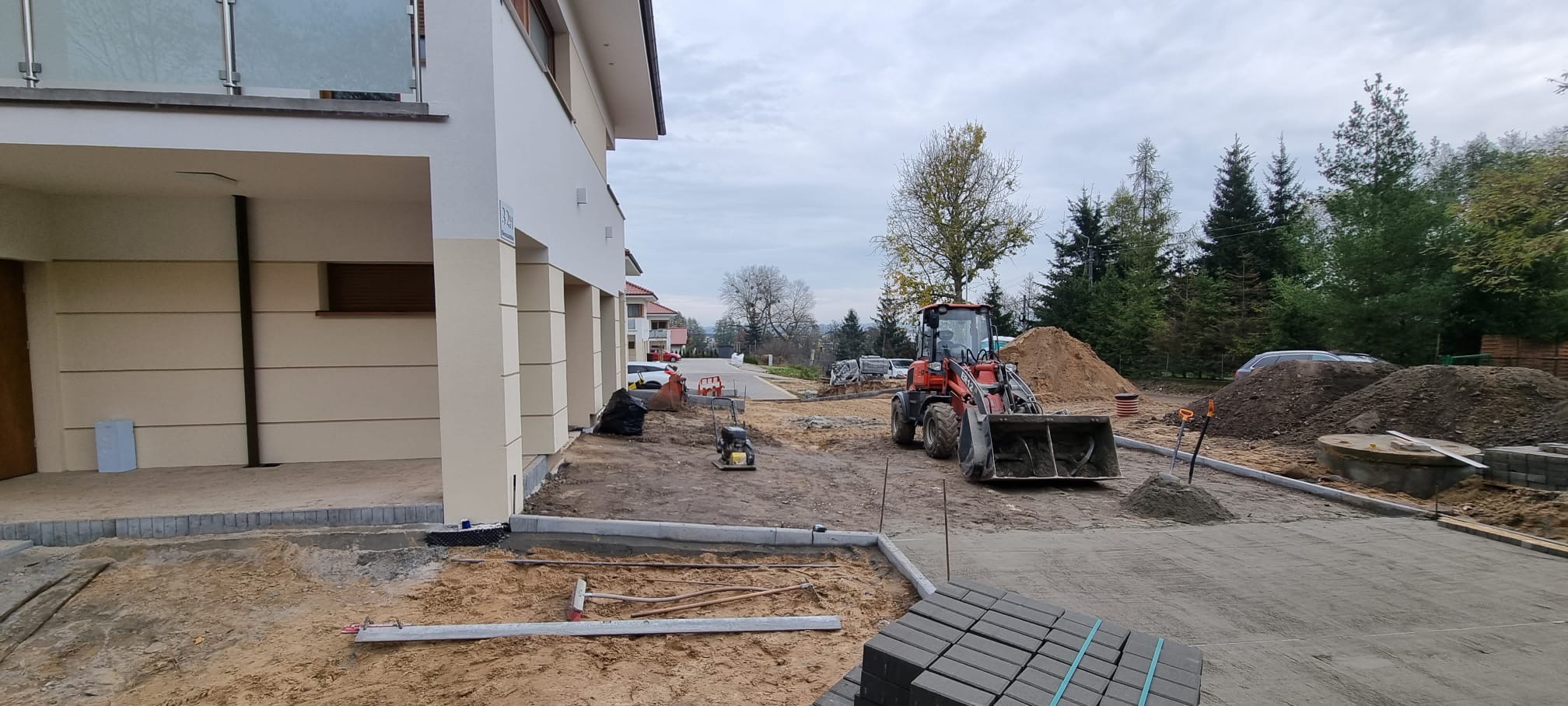 Układanie kostki brukowej przed nowo wybudowanym budynkiem, z widocznym sprzętem budowlanym i stertami ziemi w tle.
